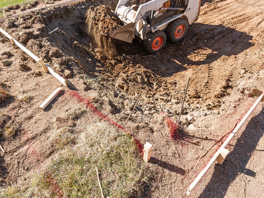 Prace ziemne przy wykopie fundamentowym z użyciem miniładowarki, widoczne drewniane szalunki i wytyczna poziomu terenu